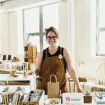 Leonora Shell, woman with brown hair and red glasses wearing a brown work apron. Standing behind a table with handmade Montessori materials at a market in Worcester, Massachusetts.